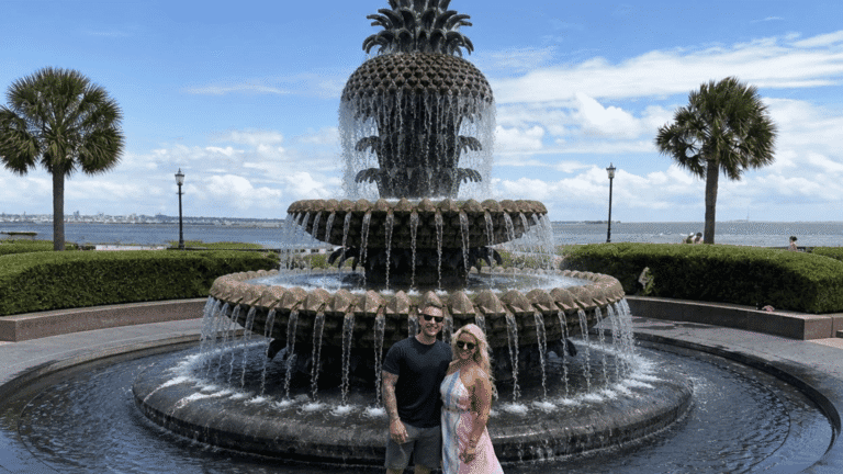 A smiling couple stands in front of a large pineapple-shaped fountain, with palm trees and the ocean in the background under a bright blue sky.