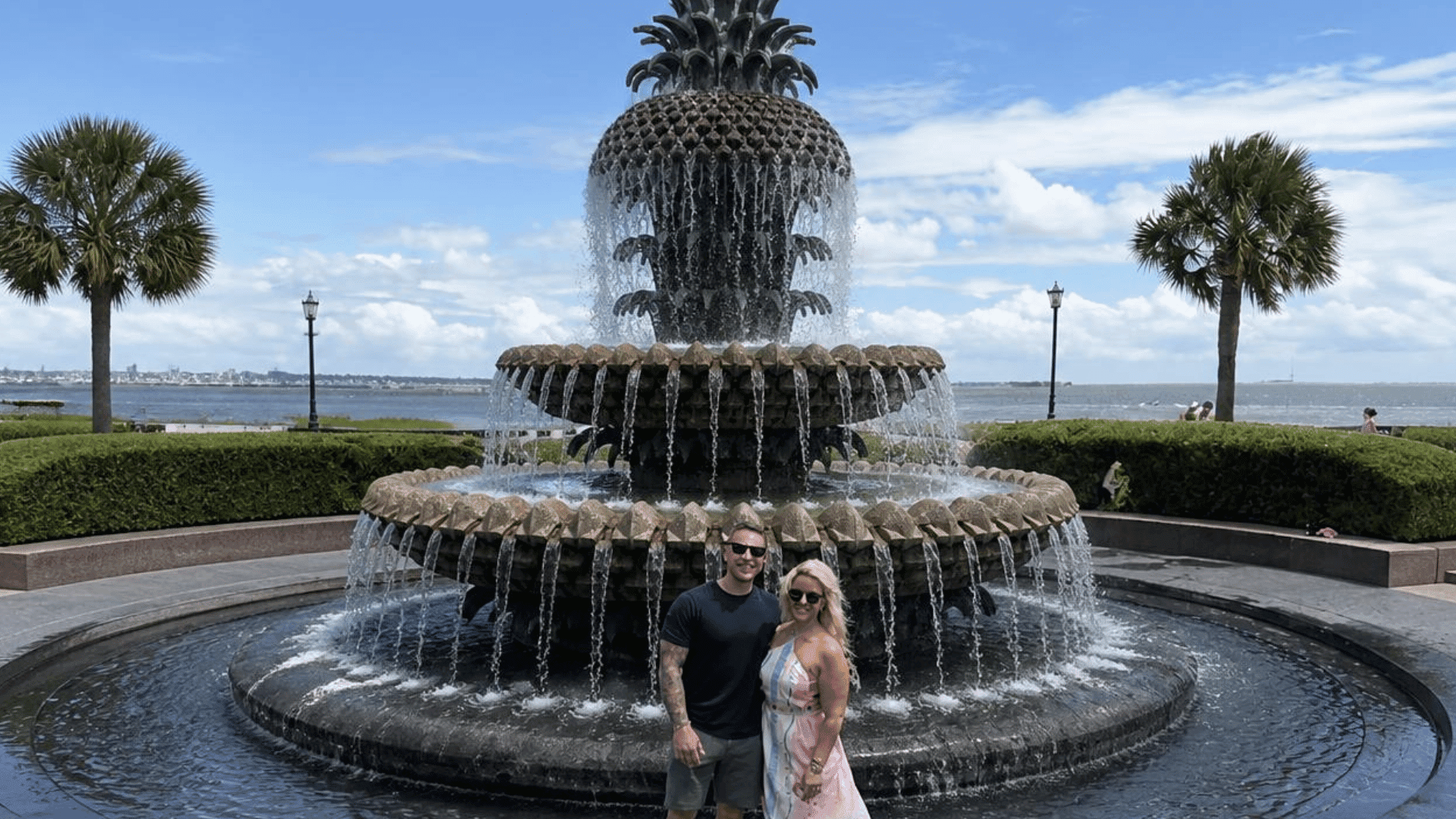 A smiling couple stands in front of a large pineapple-shaped fountain, with palm trees and the ocean in the background under a bright blue sky.