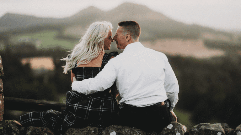 A couple sits on an ancient stone wall, embracing and gazing at each other. The backdrop features blurred green hills, creating a romantic atmosphere.