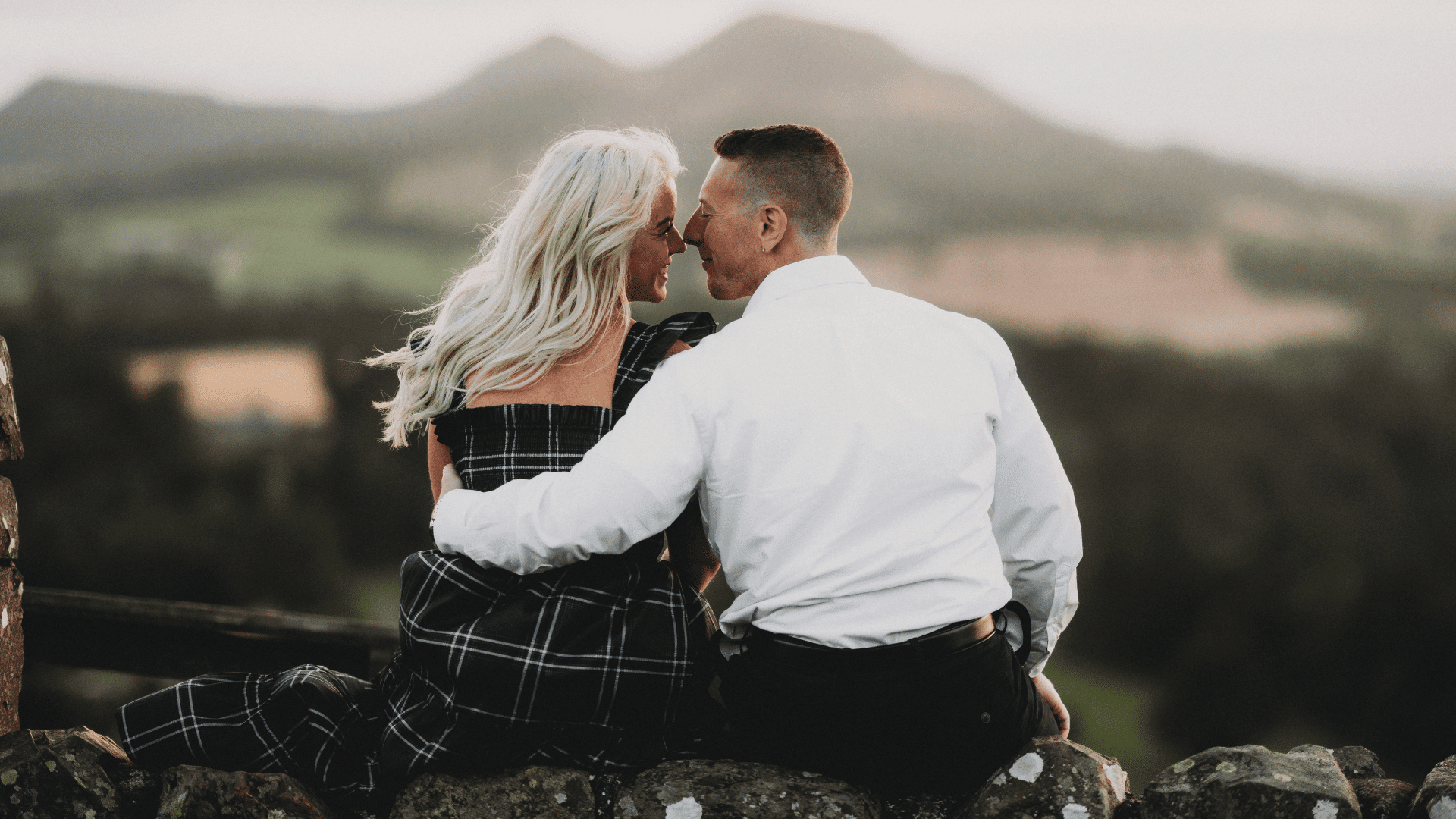 A couple sits on an ancient stone wall, embracing and gazing at each other. The backdrop features blurred green hills, creating a romantic atmosphere.