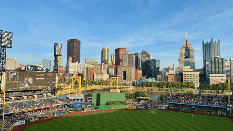 View of a baseball stadium with a game in progress, set against a city skyline under a clear blue sky. The scene is lively and bustling.