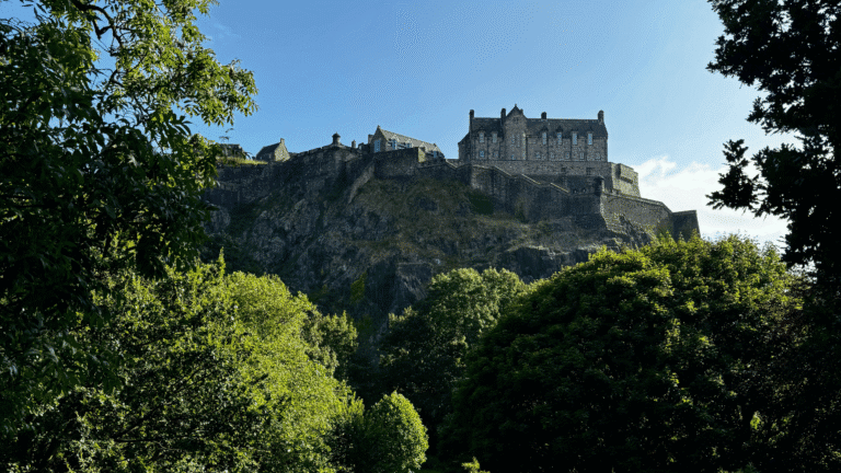 A historic stone castle sits atop a rocky hill under a blue sky, surrounded by lush green trees, conveying a sense of majesty and tranquility.