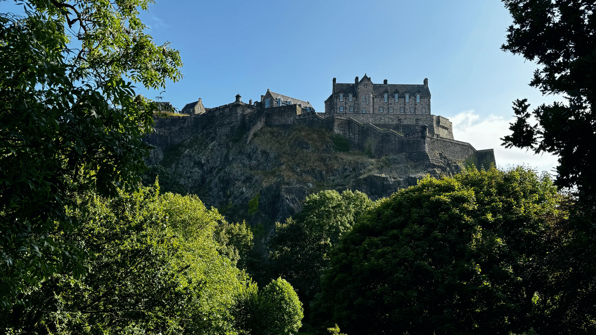 A historic stone castle sits atop a rocky hill under a blue sky, surrounded by lush green trees, conveying a sense of majesty and tranquility.