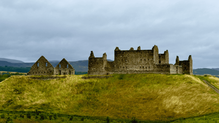 Ruthven Barracks, ancient stone ruins on a grassy hill under a cloudy sky. The large main structure is flanked by two smaller buildings, evoking a sense of history.