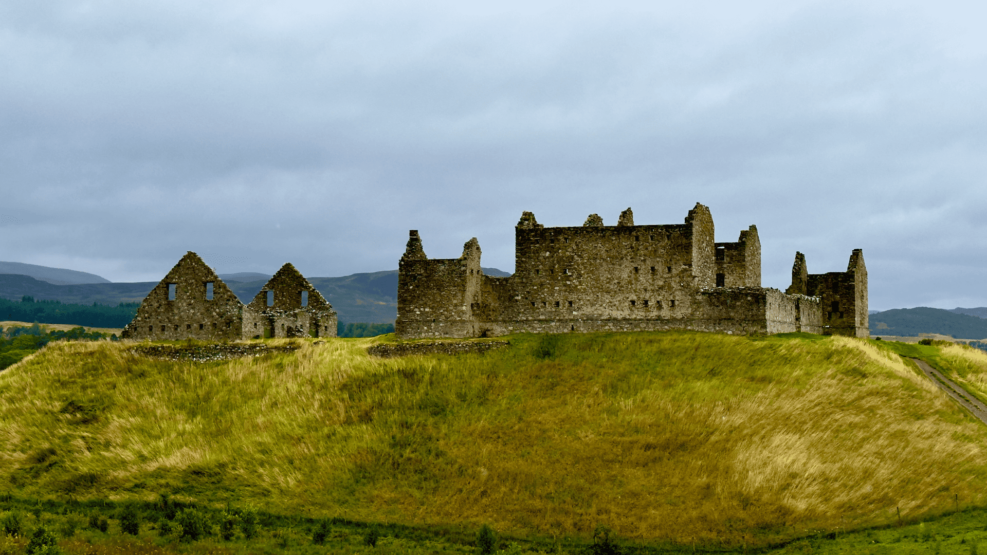 Ruthven Barracks, ancient stone ruins on a grassy hill under a cloudy sky. The large main structure is flanked by two smaller buildings, evoking a sense of history.
