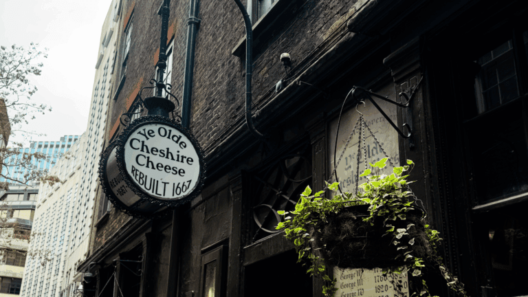 Alt text: "Historic pub sign for 'Ye Olde Cheshire Cheese, Rebuilt 1667', hangs on a dark brick building. A hanging basket with green plants adds charm. Sky and modern buildings peek through the alleyway, blending old and new."