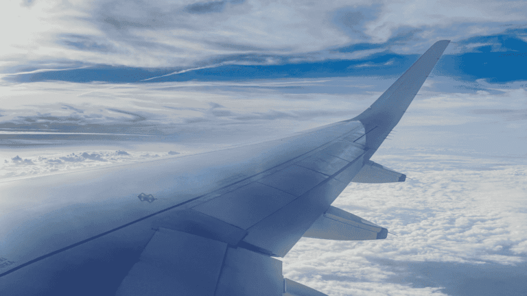 View from an airplane window showing a sleek wing against a backdrop of fluffy white clouds and a vibrant blue sky, conveying a serene and expansive feel. Used to describe a blog answering common travel questions.