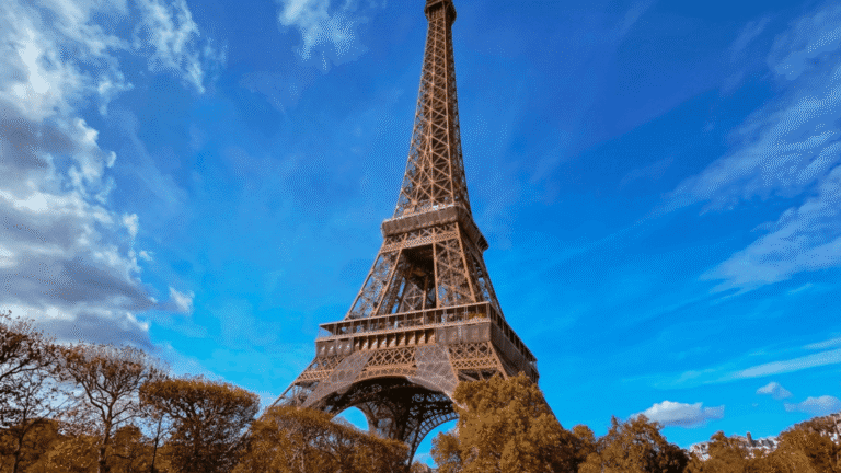 The Eiffel Tower stands against a vibrant blue sky with scattered clouds, surrounded by lush trees. The image conveys a sense of grandeur and tranquility.