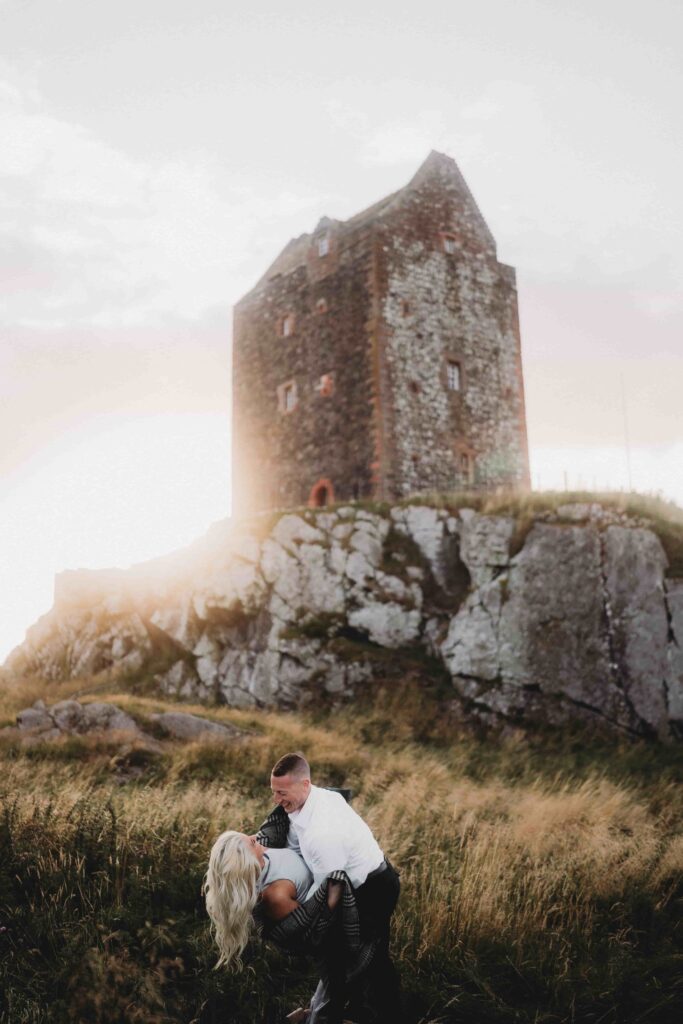 Dramatic romantic portrait of Nick and Courtney at ancient Scottish castle tower perched on rocky hilltop. Nick in white shirt dips Courtney back in intimate pose in golden windswept grass field in foreground. Courtney with long blonde hair wears plaid outfit. Behind them, imposing medieval stone tower rises from massive weathered rock outcrop against moody overcast sky with golden sunlight breaking through. Wild grassy landscape and rugged Scottish countryside surrounds them. Atmospheric engagement photography showcasing the dramatic beauty of Scotland and their deep connection to the country where their love story transformed.