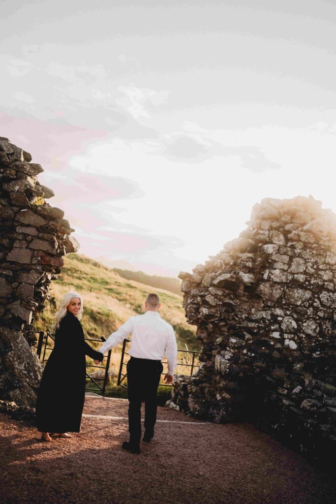 Nick and Courtney walking hand-in-hand between ancient Scottish castle ruins at golden hour sunset. Courtney in black dress with blonde hair looks back toward camera while holding Nick's hand. Nick in white dress shirt and dark pants leads her along gravel path. Towering weathered stone walls frame both sides creating dramatic corridor through castle remains. Golden sunlight streams through creating warm ethereal glow and lens flare. Rolling green Scottish hills visible in misty background. Romantic couple portrait at historic site capturing their love story in Scotland. Atmospheric moody lighting perfect for engagement photography. Ancient architecture and dramatic landscape showcase Scotland's timeless beauty.