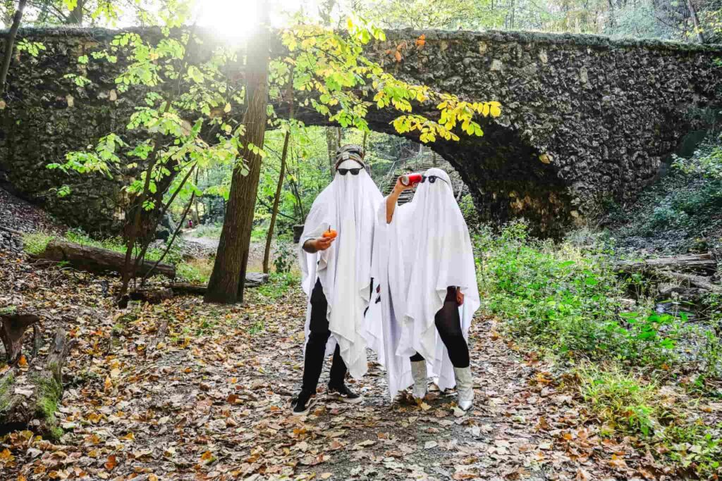 Nick and Courtney in matching ghost costumes holding drinks at historic stone ruins during fall. Both wearing classic white sheet ghost costumes with sunglasses visible through eye holes, creating playful Halloween vibe. Ancient weathered stone wall and archway structure behind them with moss and age. Forest setting with fall leaves covering ground in orange and brown. Green foliage and trees surround the ruins. Casual autumn afternoon adventure combining historic exploration with Halloween fun. Likely at Pennsylvania historic site or castle ruins.