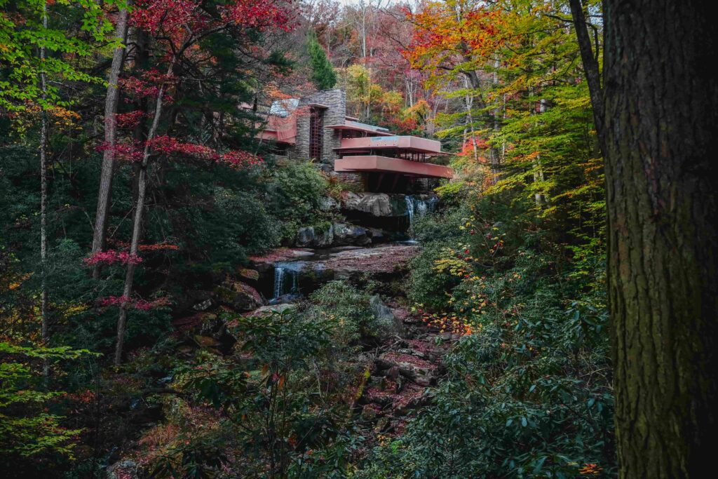 Frank Lloyd Wright's iconic Fallingwater house nestled in Pennsylvania forest during peak autumn. The famous modernist home features distinctive horizontal cantilevered terraces in earthy red-brown tones extending over natural waterfall. Stone chimney anchors the structure vertically. House integrated seamlessly with surrounding nature as cascading waterfall flows beneath and around the building over natural rock formations. Vibrant fall foliage surrounds the scene with brilliant red, orange, yellow, and green leaves. Dense forest provides natural privacy. Multiple tiers of waterfalls visible in foreground with moss-covered rocks. Architectural masterpiece demonstrating organic architecture principles blending human design with natural landscape. Mill Run, Pennsylvania.