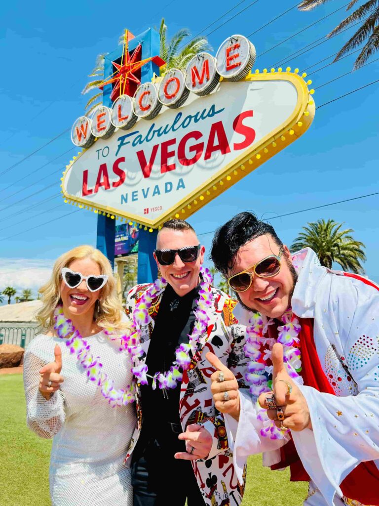 Nick and Courtney posing with Elvis Presley impersonator at the iconic 'Welcome to Fabulous Las Vegas Nevada' sign. All three wearing oversized novelty sunglasses and colorful Hawaiian lei necklaces, giving enthusiastic thumbs up and shaka hand gestures. Courtney on left in white outfit with heart-shaped sunglasses. Nick in center wearing black and white patterned shirt with polka dot sunglasses. Elvis impersonator on right in classic white rhinestone-studded jumpsuit with red cape and gold aviator sunglasses. The famous retro sign towers behind them featuring red 'WELCOME' letters, diamond shapes, starburst, and yellow border with light bulbs. Bright blue sky, palm trees, and clear desert sunshine. Classic Las Vegas tourist photo capturing the fun, kitschy energy of the city.