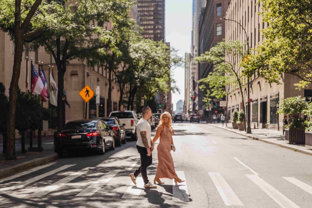 Nick and Courtney crossing downtown New York City street hand-in-hand in romantic candid moment. Courtney with blonde hair wearing elegant blush pink maxi dress and heeled sandals looking back over shoulder. Nick in white t-shirt and black pants showing tattooed arms walking beside her. Tree-lined urban street with parked cars, tall office buildings, pedestrian crosswalk, traffic lights, and American flags visible. Dappled sunlight creates beautiful shadows across pavement. Green summer trees frame the scene. Classic city couple portrait capturing authentic connection in their hometown. Professional engagement or lifestyle photography in downtown New York City, New York.