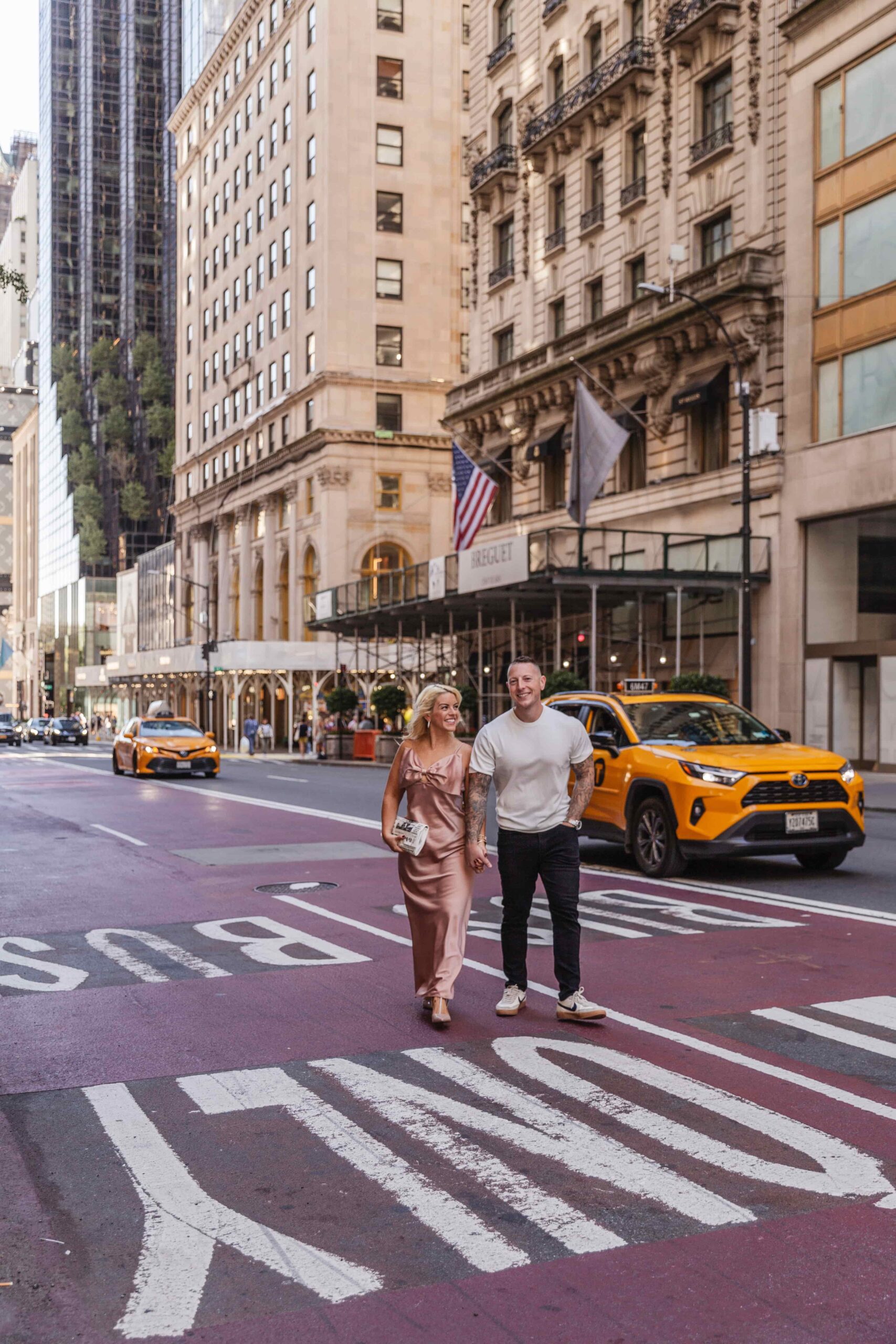 A couple walks hand in hand on a city street with historic buildings. Yellow taxis drive by, creating a lively urban atmosphere.
