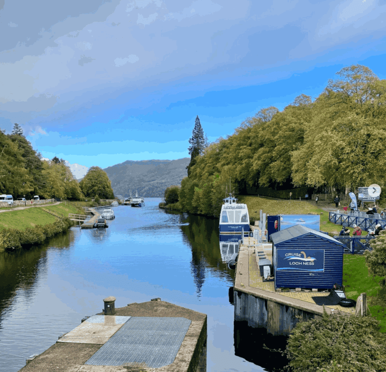Scenic view of Caledonian Canal at Loch Ness, Scotland showing cruise boats docked along peaceful waterway. Blue 'Cruise Loch Ness' ticket booth on wooden dock with tour boats moored nearby. Lush green trees line both sides of calm canal reflecting in still water. Scottish Highlands mountains visible in distance under blue sky. Popular couples travel destination for budget-friendly Scotland itineraries. Honest travel guide location showcasing accessible Scottish adventure without rental car. Perfect for couple travelers exploring Scottish Highlands on limited vacation time.