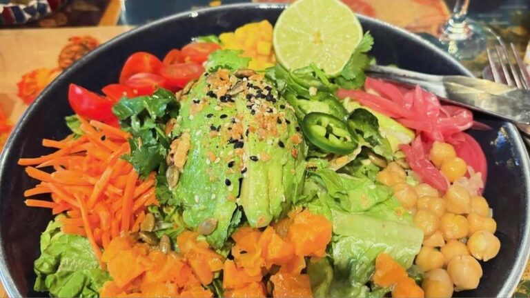 A vibrant salad bowl with sliced avocado topped with seeds, surrounded by cherry tomatoes, carrots, chickpeas, jalapeños, lime, and cilantro.