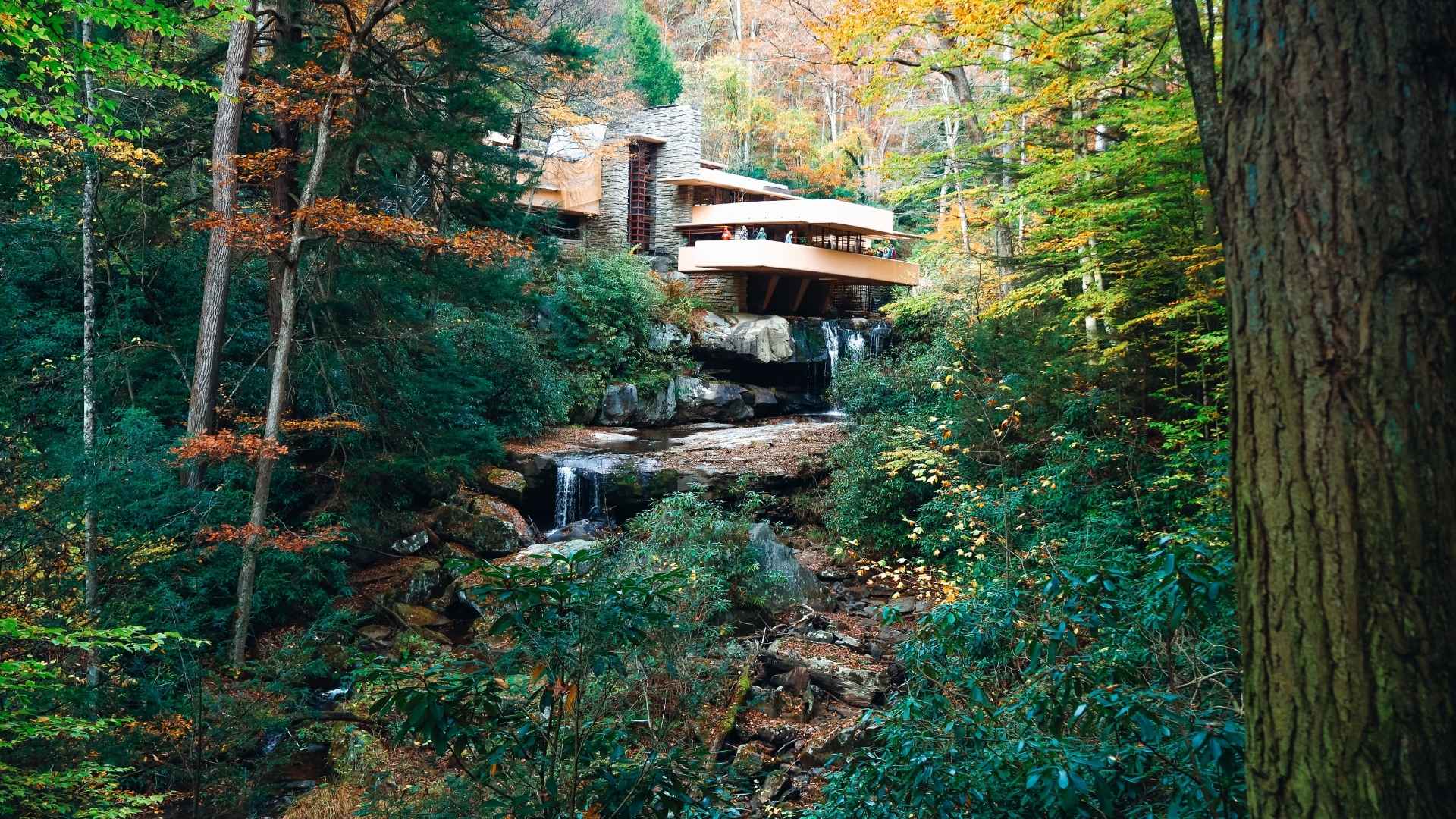 A modern house, known as Fallingwater, is perched over a waterfall amidst a lush autumn forest. The scene conveys harmony and tranquility.