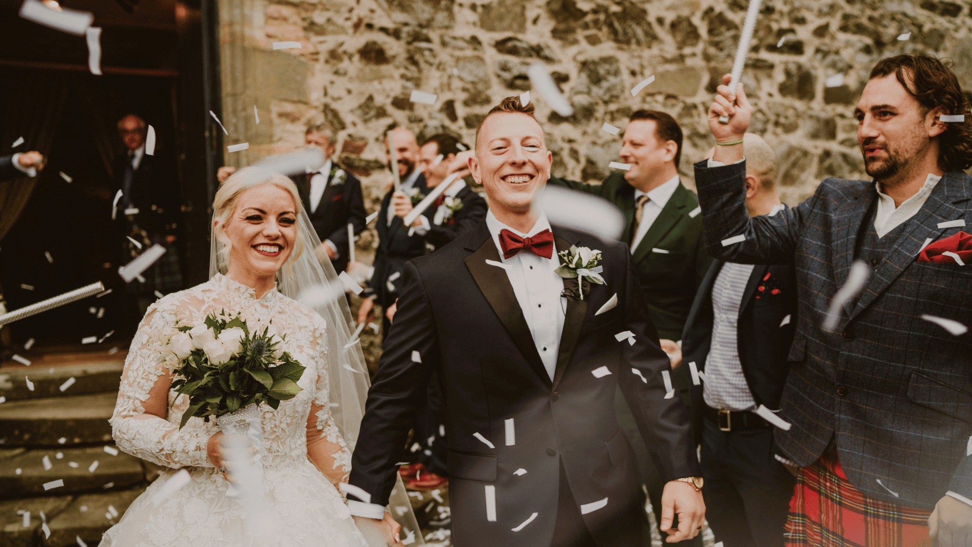 Bride and groom, smiling, walk down an aisle while guests throw confetti. The bride holds a bouquet; the scene feels joyful and celebratory.