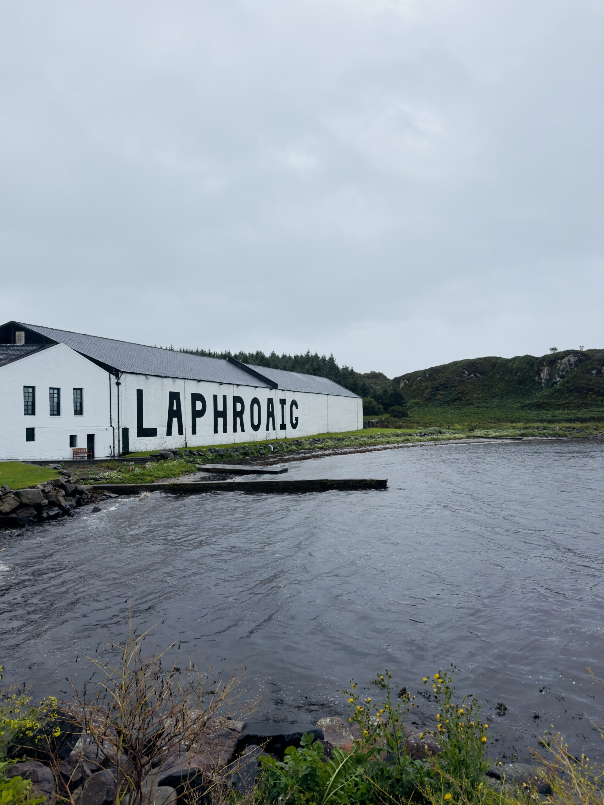 Laphroaig Distillery on the Isle of Islay, Scotland. The iconic white whisky distillery building with "Laphroaig" in large black letters overlooks a calm bay on the Islay whisky trail under overcast Scottish skies.