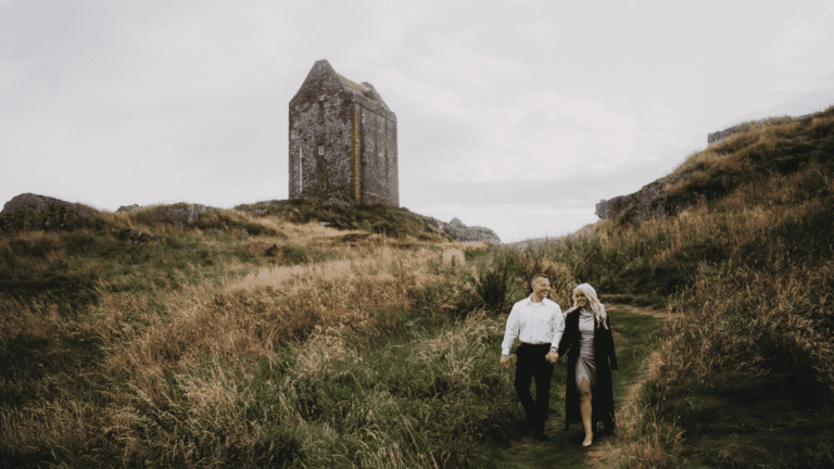 A couple strolls hand in hand on a grassy path through a hilly landscape. An old stone tower stands in the background, evoking a serene, timeless atmosphere.