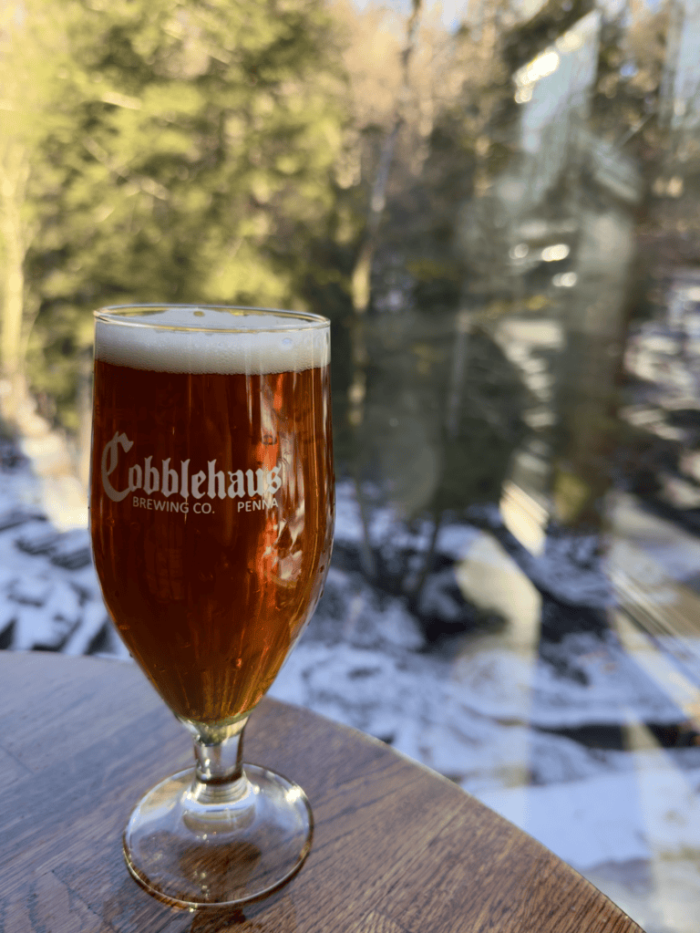 Amber beer in a stemmed Cobblehaus glass on a wooden table by the window at Cobblehaus at the Falls in Mercer, PA, with snow and bare winter trees outside.