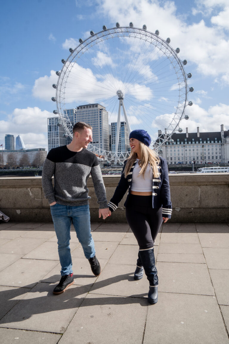 Couple walking hand in hand along the Thames with the London Eye and a bright blue sky behind them