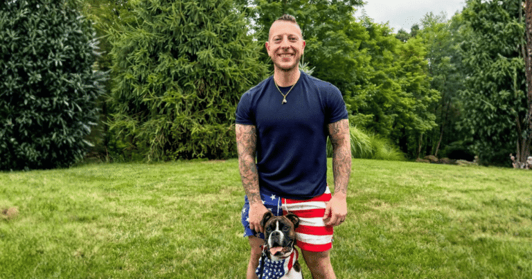 Man in American flag shorts smiling on a green lawn with a boxer dog wearing a stars and stripes bandana