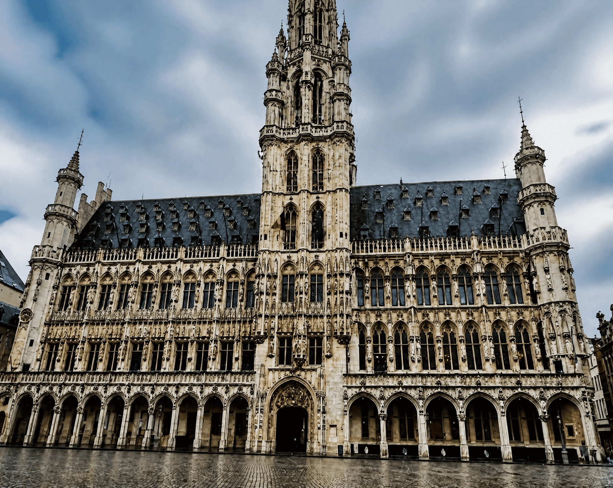 Brussels Town Hall on Grand Place, a Gothic building with an ornate stone facade and a tall central spire, under a cloudy sky.